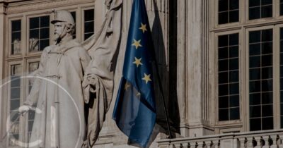 European Union flag next to a historic statue in front of a classical building facade, symbolizing EU governance or citizenship