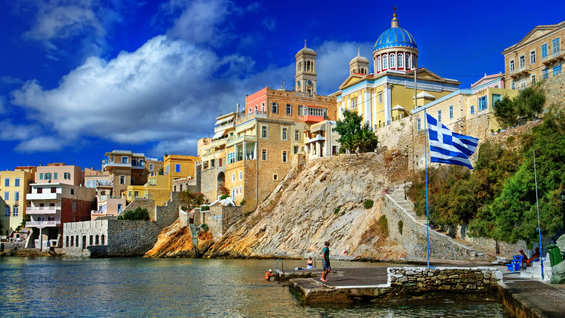 Greece Golden Visa Citizenship: How It Works in 2026 1 A coastal Greek village built on a rocky cliffside, showcasing historic stone buildings, a blue-domed church, and the Greek flag flying over a stone dock with clear blue water below. (Greek Golden Visa Citizenship)
