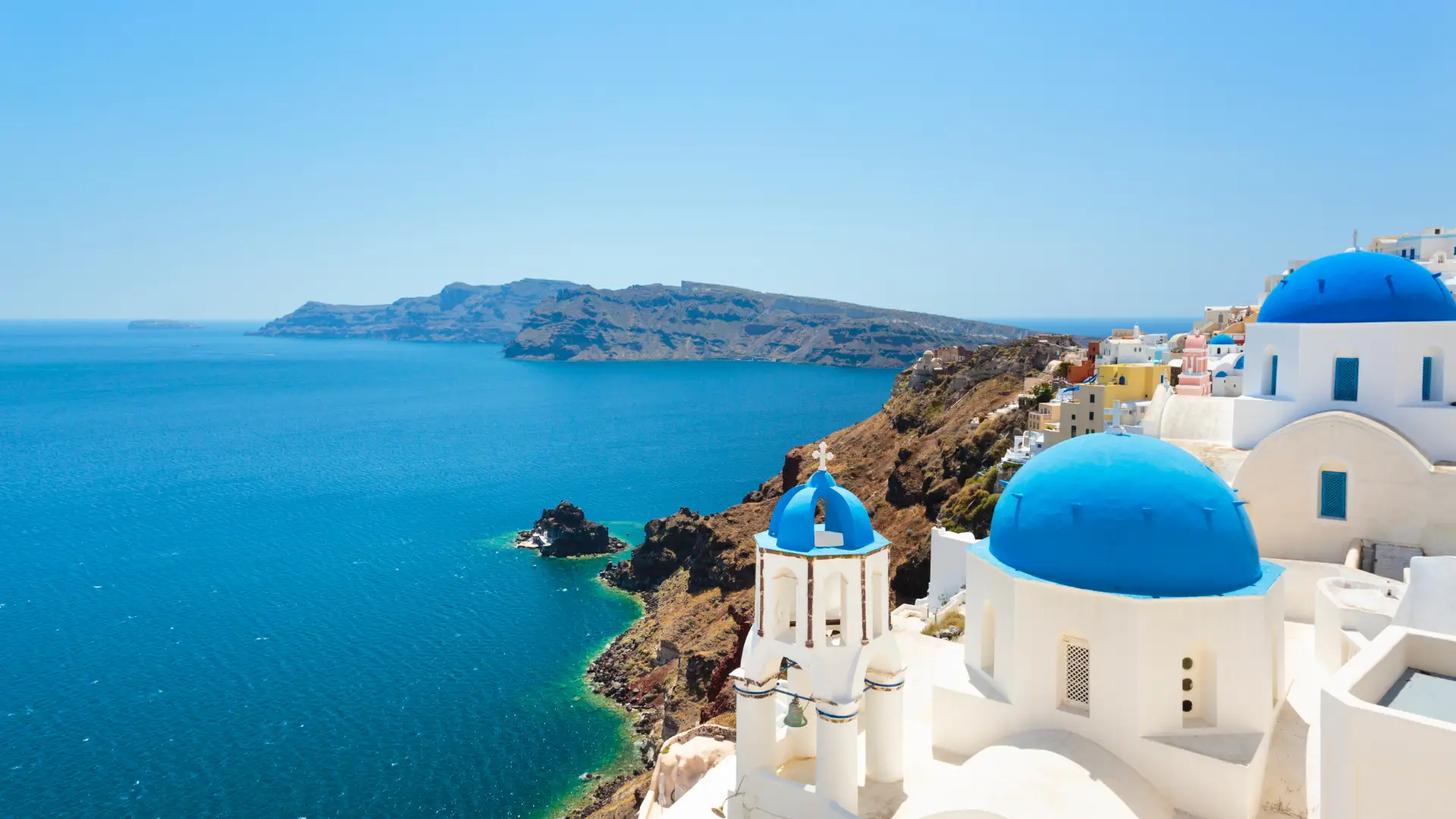 Greece Golden Visa Citizenship: How It Works in 2026 2 High-angle shot of white-washed buildings and iconic blue-domed churches in Oia, Santorini, set against volcanic cliffs and clear turquoise sea under a bright blue sky. (Greek Golden Visa Citizenship)